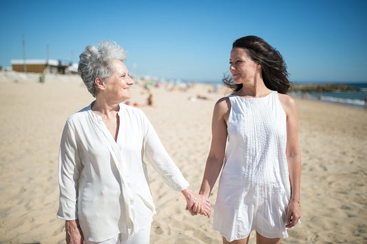 An older adult woman holds hands with her adult daughter while they walk on the beach.
