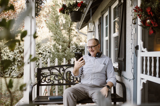 An older man smiles at his phone while sitting on a porch.