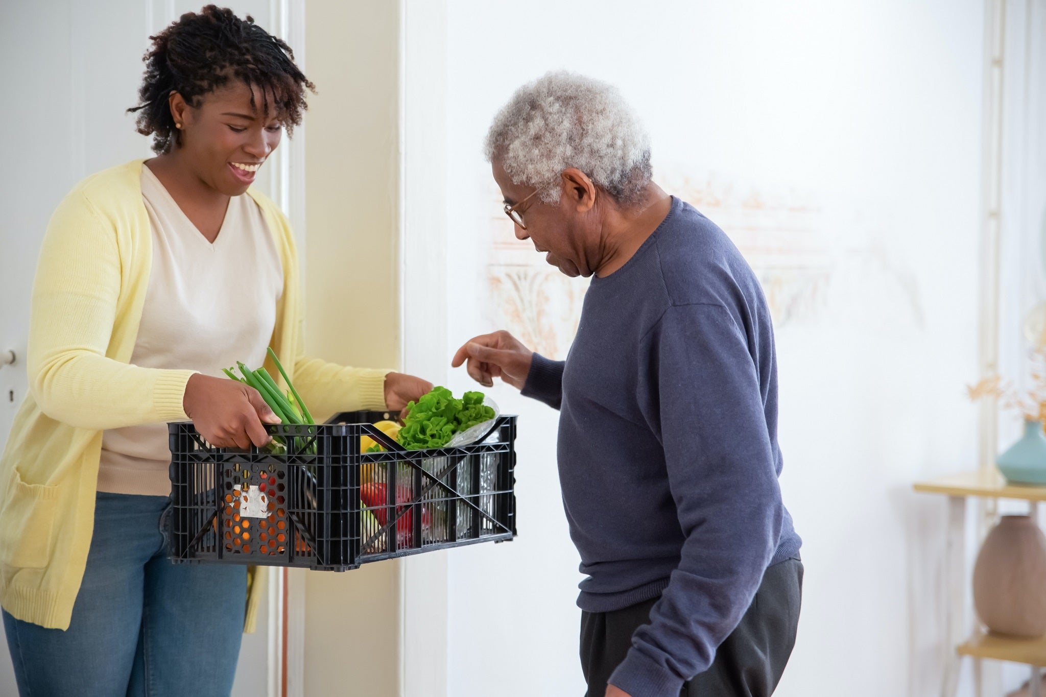 An adult woman holds a basket of groceries for an elderly man. 
