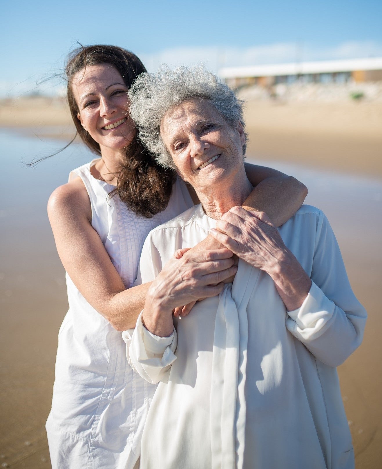 An adult woman lovingly embraces an elderly woman from behind while standing on a beach. 