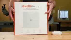 A man unboxes a weight scale, sets it on a table, then presses on the scale with his hands to obtain an example weight.